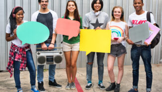 Groep diverse jongeren staat naast elkaar tegen een metalen wand en houdt grote gekleurde tekstballonnen vast, één jongen draagt een retro radio en anderen hebben koptelefoons om hun nek.